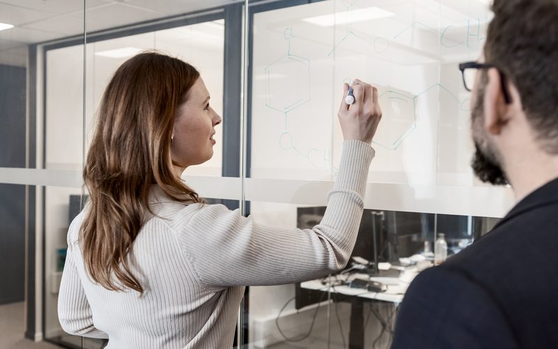 Colleague drawing a chemical structure on a glass board while a teammate watches